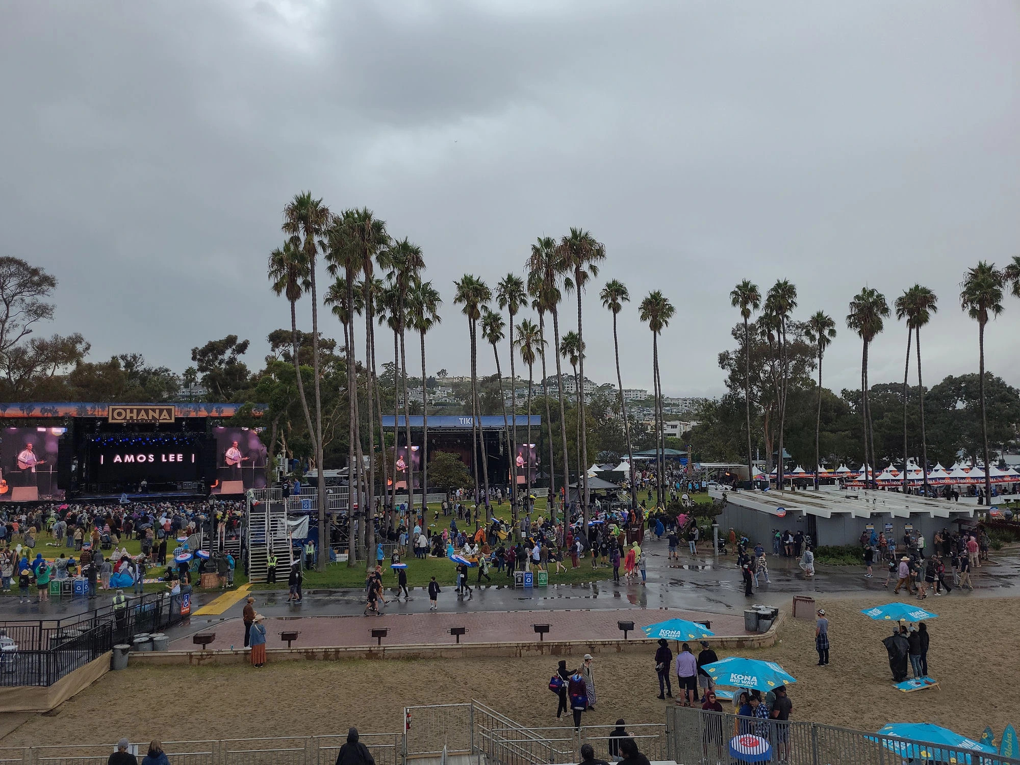 Ohana Festival main stage at Doheny State Beach under storm-gray skies, Amos Lee on the screens, rain ponchos in the crowd, 2023
